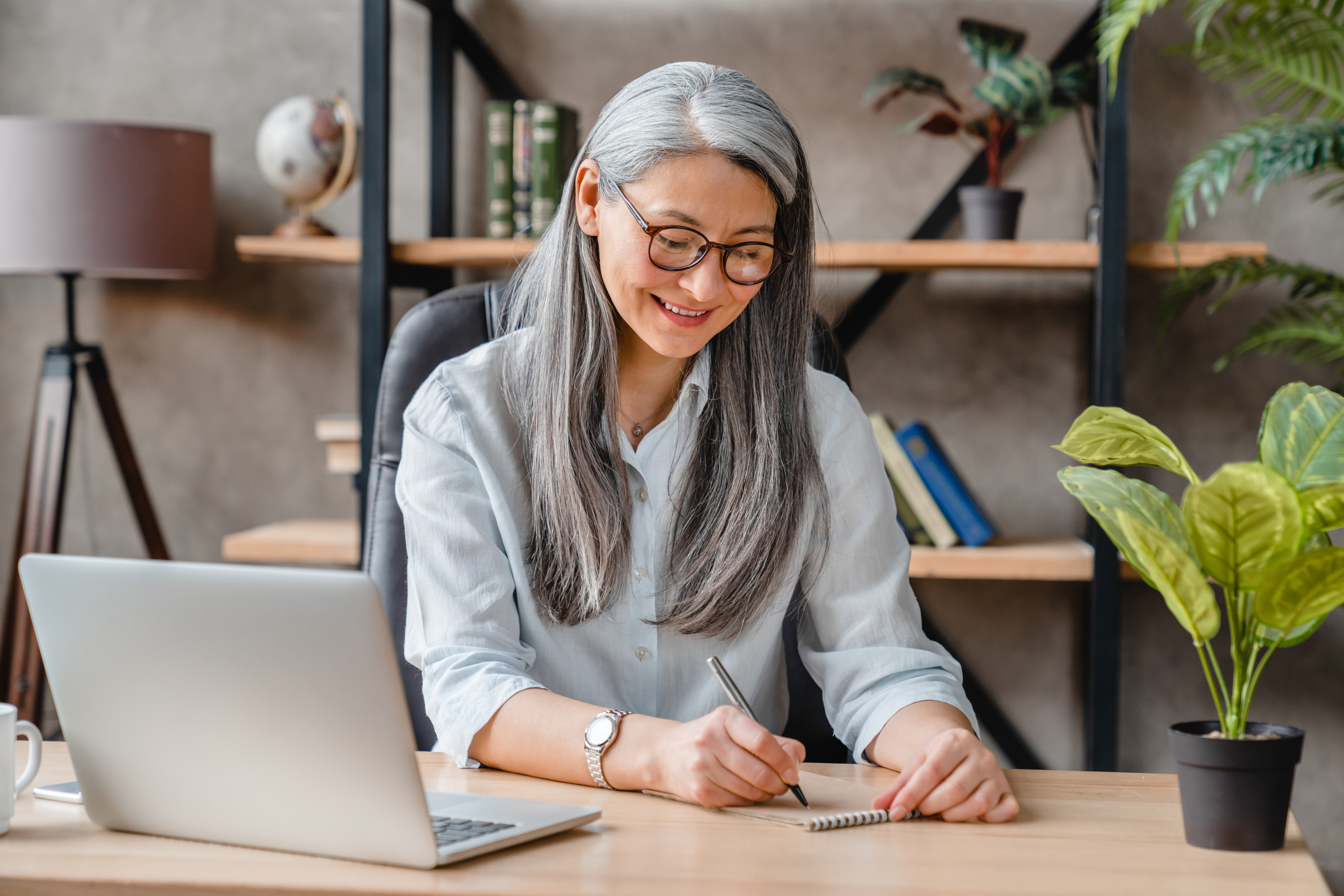 Beautiful middle-aged woman with grey hair writing on her desk in office A woman sits at a desk while writing something down in a small notebook.