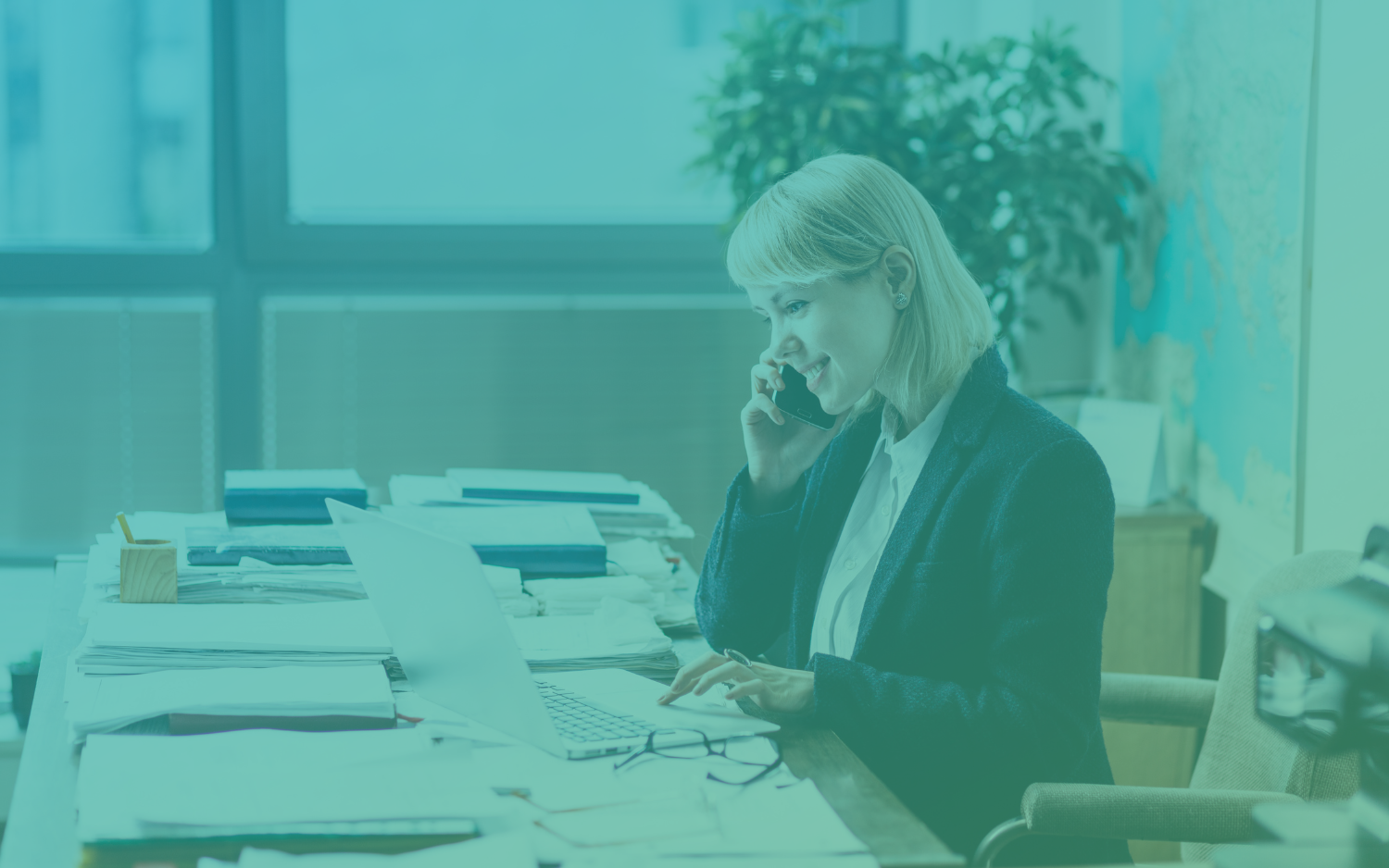 Modern Guide to PR A person with short blonde hair is sitting at a desk on the phone looking at a laptop, and they are surrounded by stacks of paper.